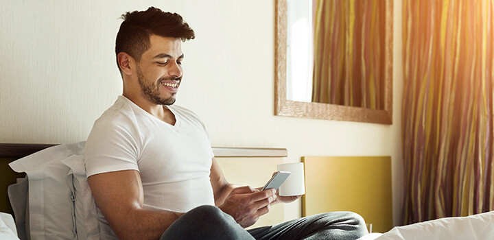 マグカップを片手に笑顔でスマートフォンに視線を落とす男性の画像