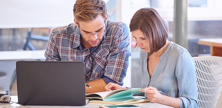 デスクの上でノートパソコンを開き、その横に置かれた書類を真剣に見ている男女の画像