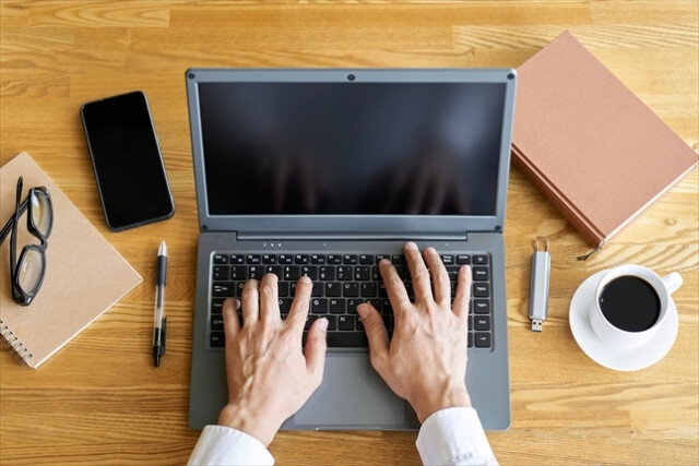 ノートパソコンと仕事道具が置かれたデスクで作業をする男性の手元